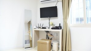 A room with a white air conditioner, a flat-screen TV, a desk with various electronic devices, a mirror, and a window with curtains. - JW STAY in HONGDAE - Hostel (Seoul)