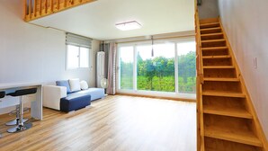 Une pièce spacieuse avec un escalier en bois, un canapé, une table à manger avec des chaises et de grandes fenêtres donnant sur un paysage verdoyant.