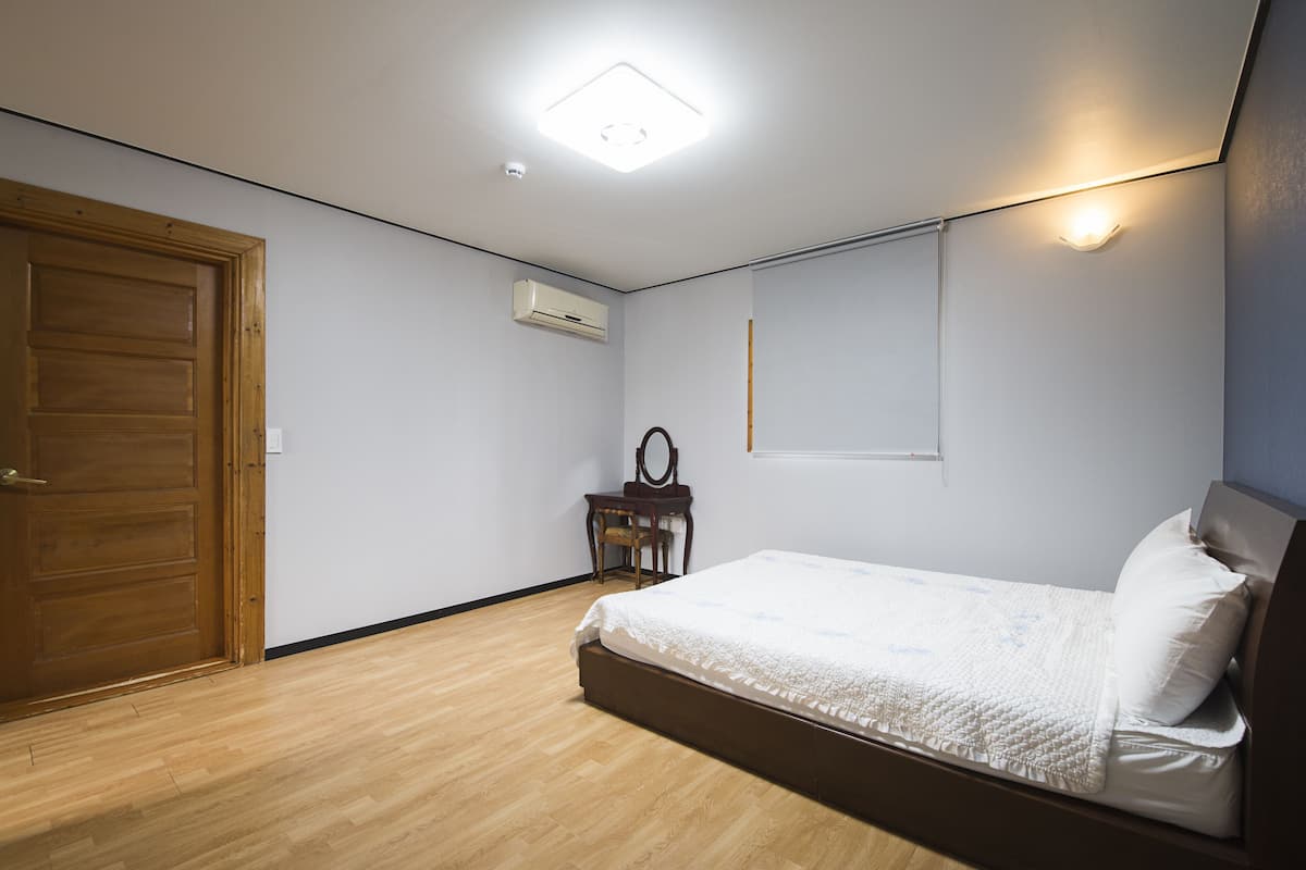 A bedroom with a wooden door, a bed with white bedding, a small table with a mirror, and a ceiling light.
