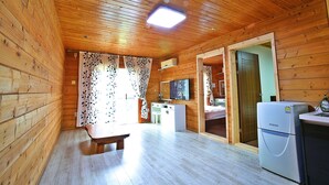 L’intérieur d’une cabane en bois, avec un plancher en bois, un banc, un bureau blanc, un réfrigérateur et une porte donnant sur une autre pièce.