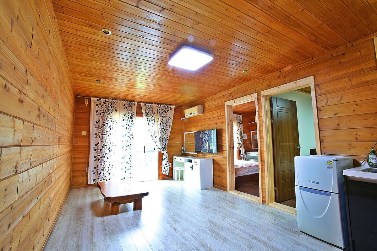 A wooden cabin interior with a wooden floor, a bench, a white desk, a refrigerator, and a door leading to another room.