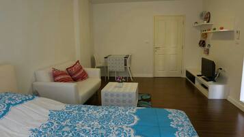 A small, well-lit room with a bed, a sofa, a small table with a vase, a television, and a door leading to another room.