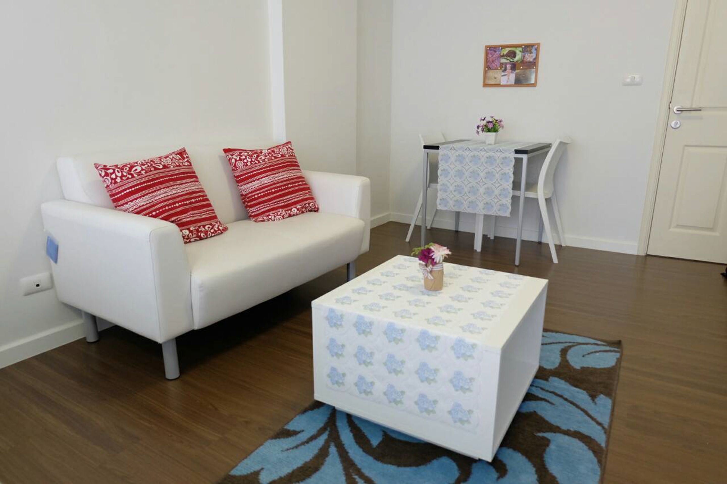 A white sofa with red patterned pillows, a small table with a floral tablecloth, and a blue patterned rug.