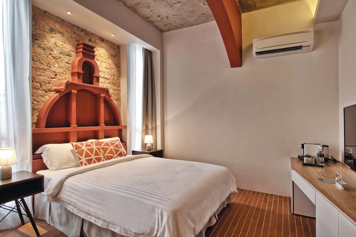 Un dormitorio con pared de ladrillo, cabecera de madera, cama con ropa de cama blanca, mesita de noche con lámpara y cómoda de madera con cafetera.