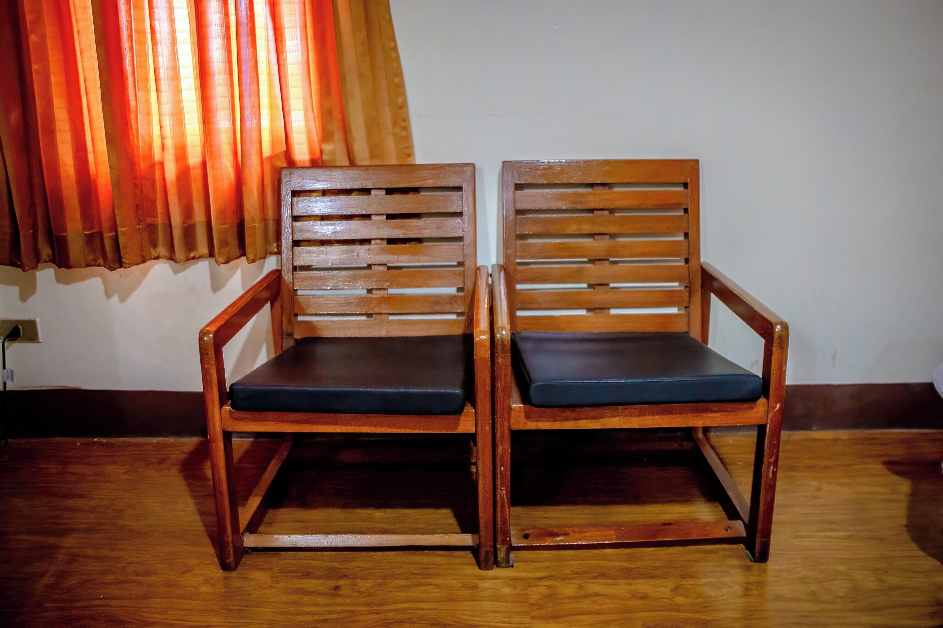 Two wooden chairs with dark cushions against a wall with a curtain.