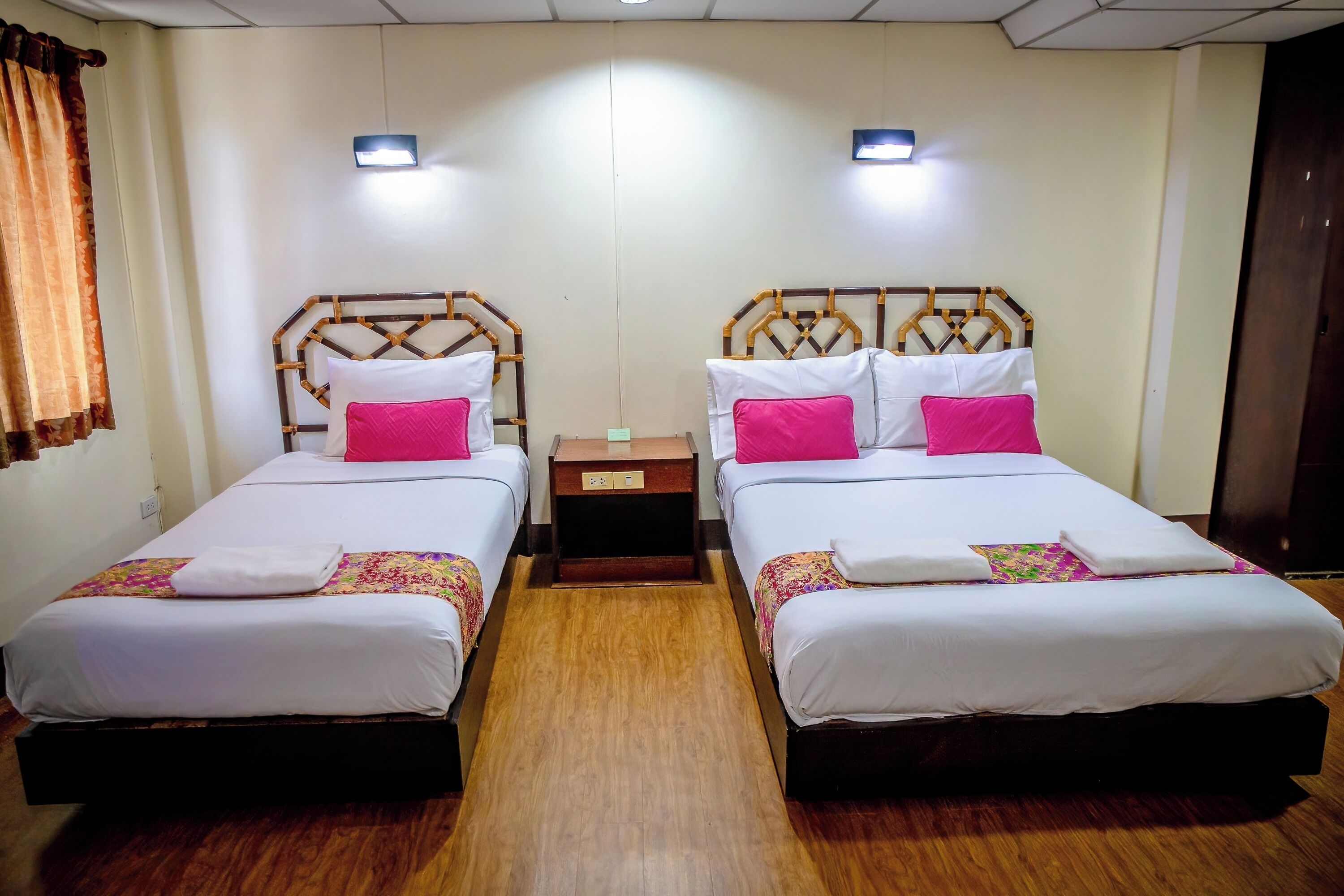 Habitación de hotel con dos camas individuales, cada una con cabecero de madera, ropa de cama blanca y almohadas rosas. Hay una mesita de noche de madera entre las camas y el piso es de madera.