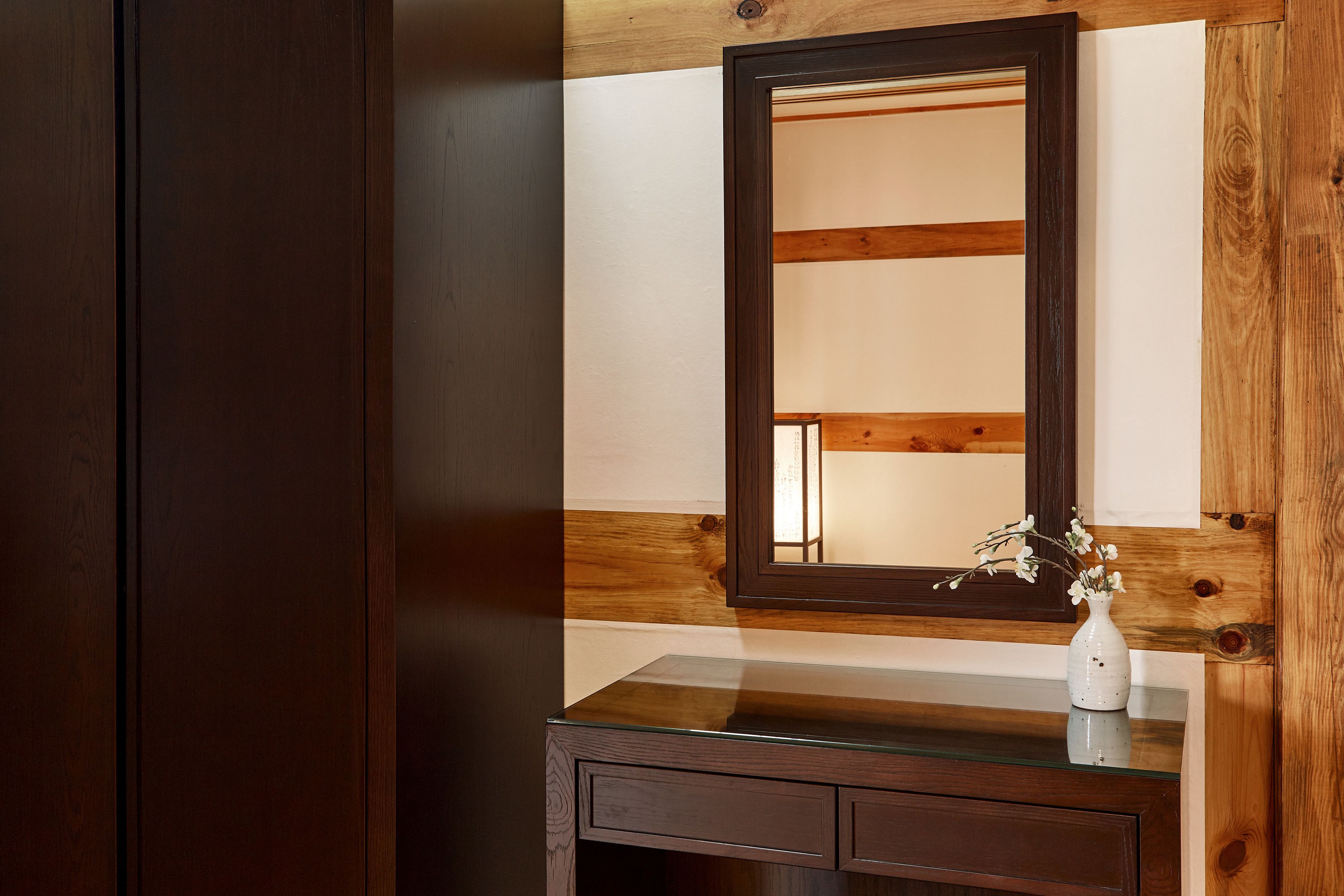 A wooden vanity with a mirror, a vase with flowers, and a glass countertop.