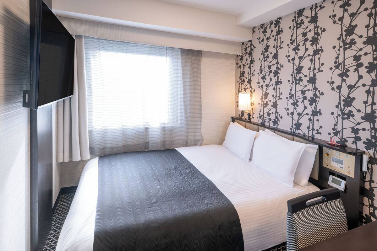 Une chambre d’hôtel avec un grand lit, une table de chevet avec une lampe, un téléviseur fixé au mur et une fenêtre avec des rideaux.