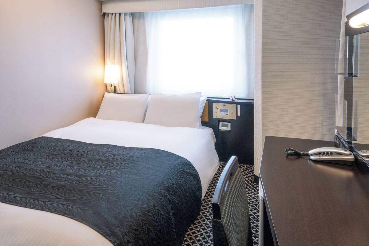 Une chambre d’hôtel avec un lit, un bureau avec un téléphone, un panneau de commande et une fenêtre avec des rideaux.