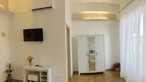 Quarto com uma secretária branca, televisão de tela plana na parede, guarda-roupa e uma janela grande com cortinas leves.