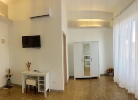 Quarto com uma secretária branca, televisão de tela plana na parede, guarda-roupa e uma janela grande com cortinas leves.
