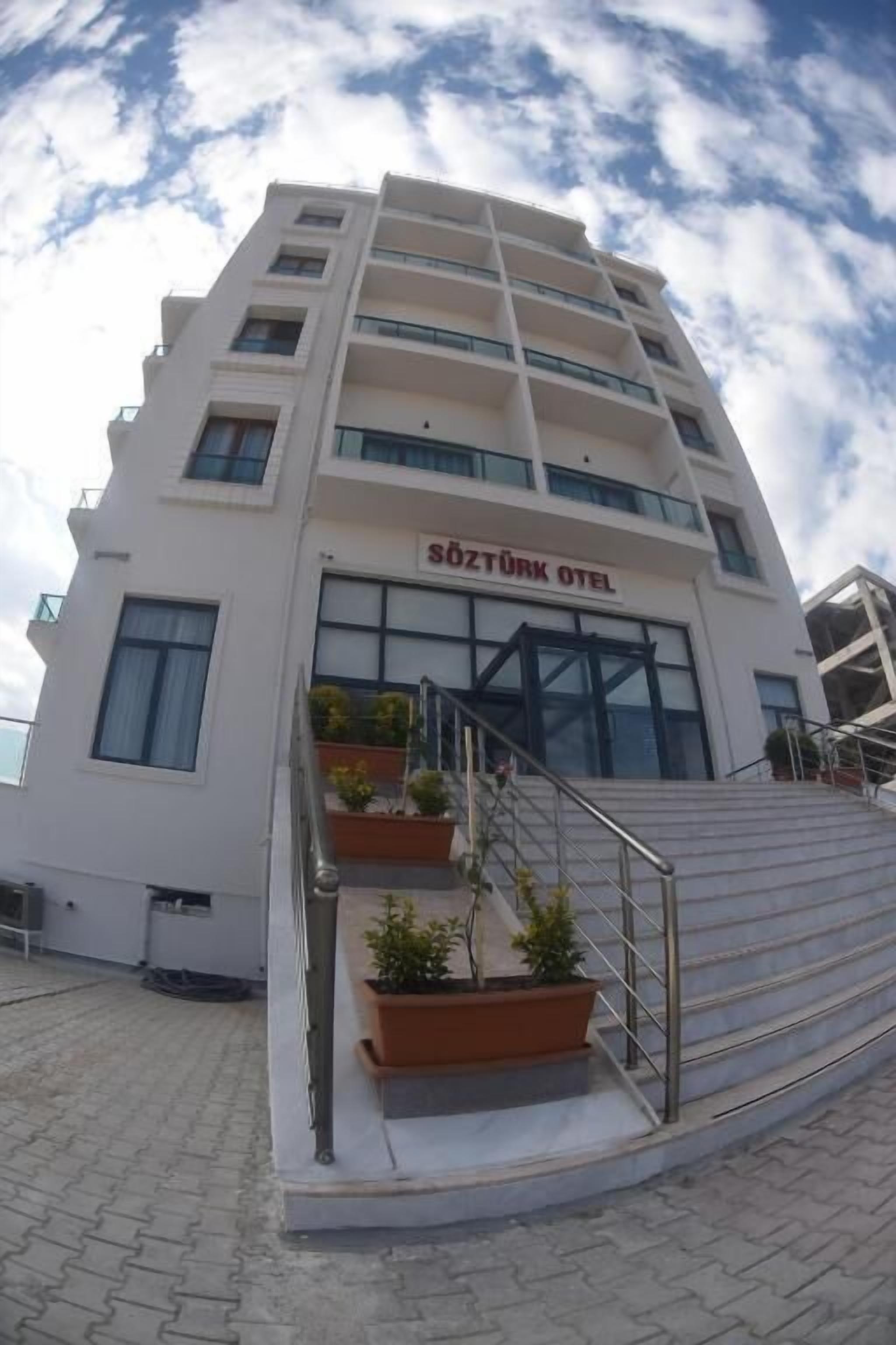 Un moderno edificio de hotel con un cartel que dice "SOZTÜRK OTEL". Tiene varios pisos, una escalera que conduce a la entrada y macetas exteriores con plantas.