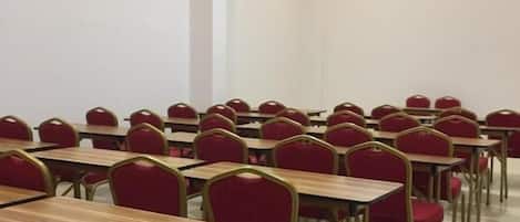 Un salón de conferencias con filas de mesas y sillas, paredes blancas y luces en el techo.
