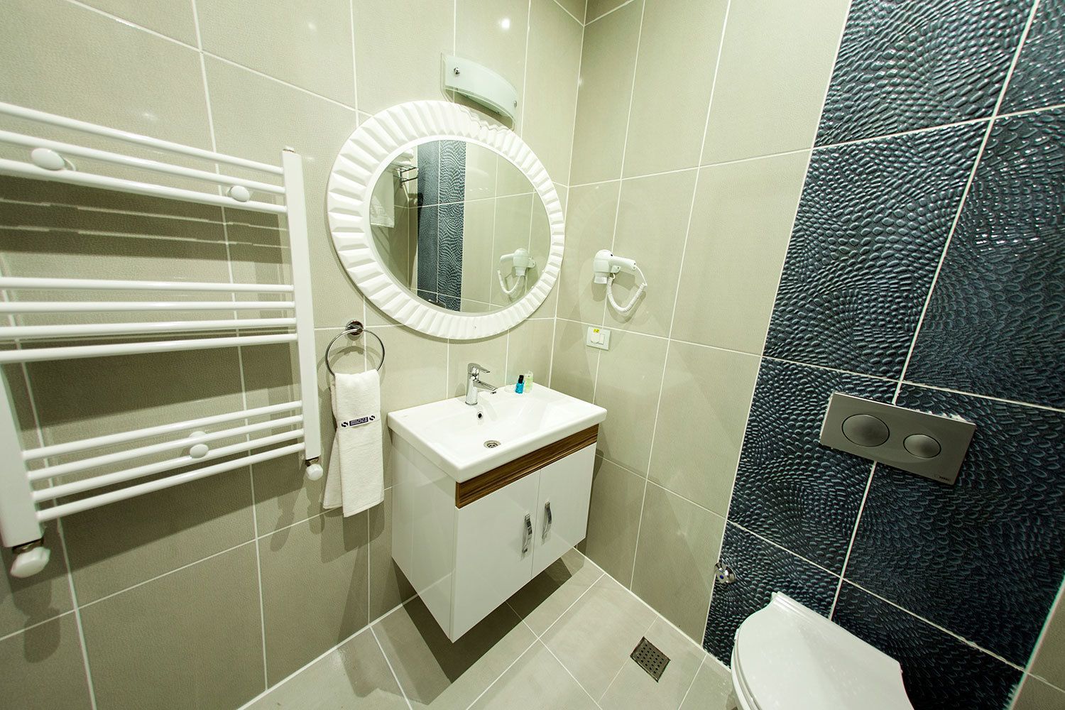 A modern bathroom with a white vanity, a round mirror, a towel rack, and a toilet.