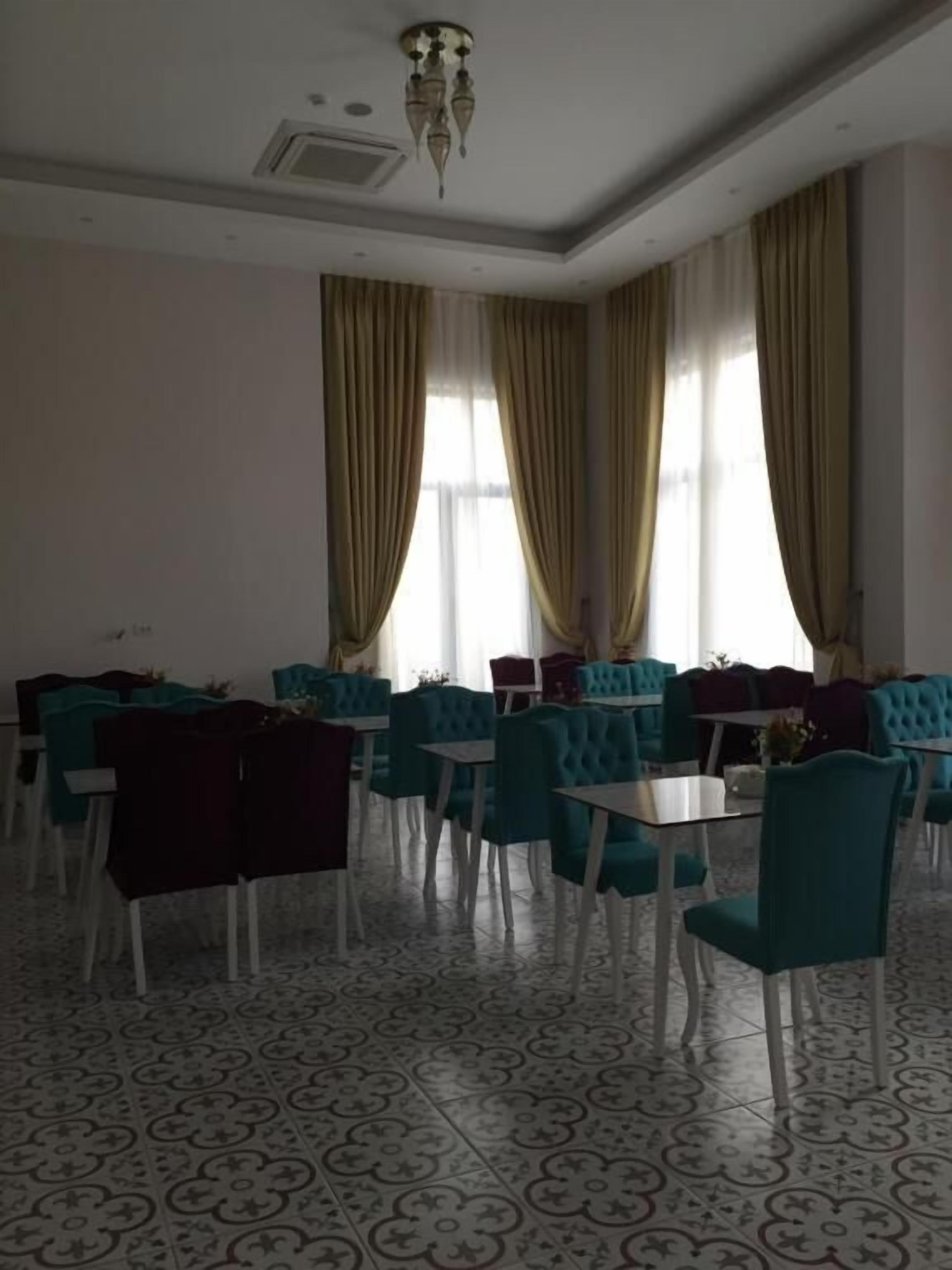 Un comedor con sillas tapizadas en color aguamarina y mesas, ventanas amplias con cortinas doradas y un suelo estampado.