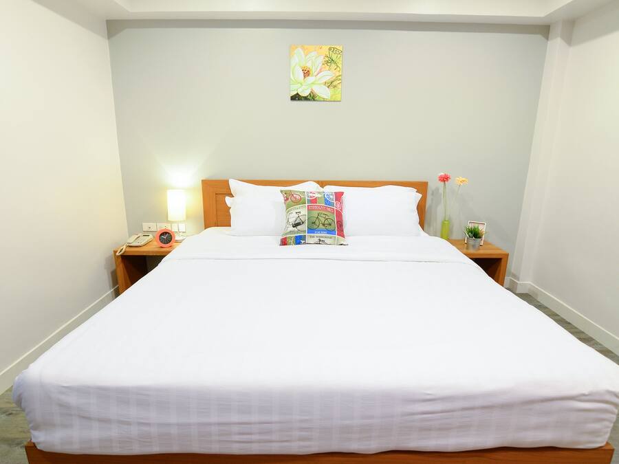 A neatly made bed with white bedding, a bedside table with a lamp, a clock, and a small vase with flowers.