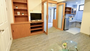 A compact living space with a wooden entertainment unit, a glass coffee table, and a kitchenette area. - BJ Boutique (Rayong)