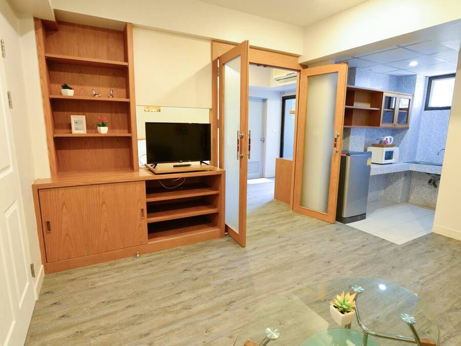 A compact living space with a wooden entertainment unit, a glass coffee table, and a kitchenette area.