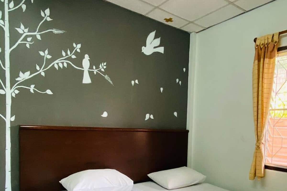 Hotelkamer met een bed, twee kussens, een hoofdbord, een raam met gordijnen en een wanddecoratie van een boom en vogels.