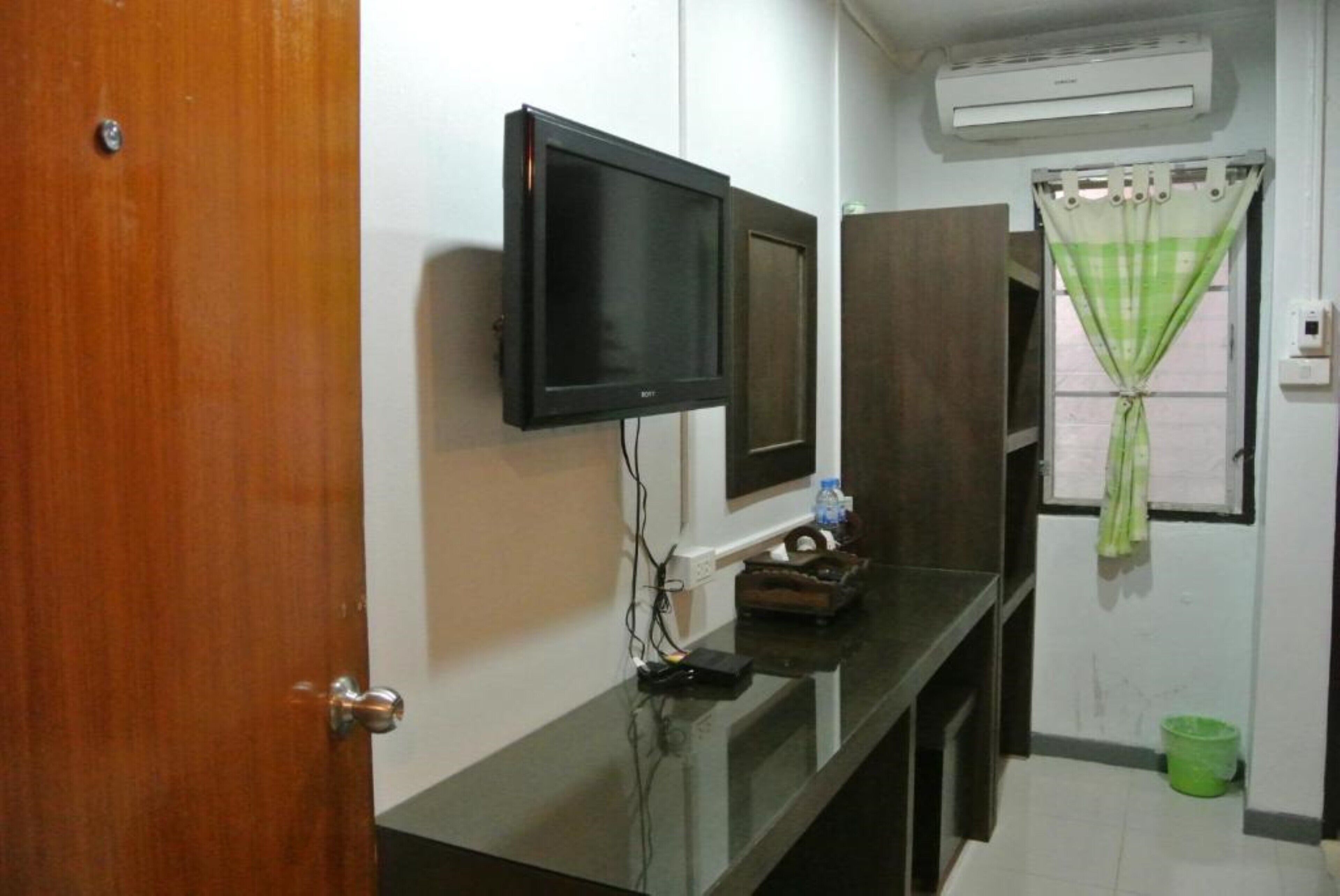 A room with a wooden door, a black TV on the wall, a window with green curtains, and an air conditioner above the window.