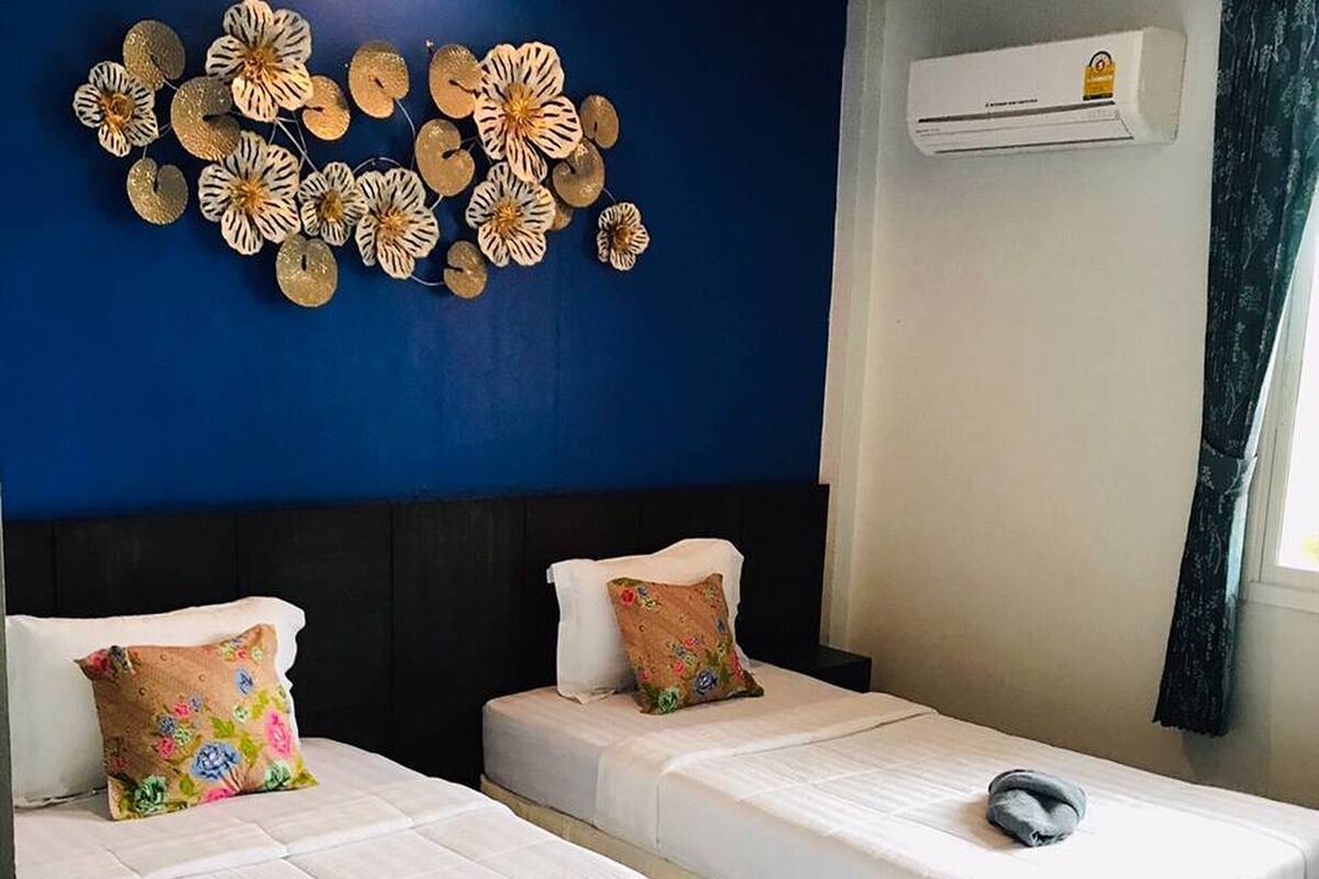 Een hotelkamer met twee bedden, elk voorzien van een witte spreidek en een geplooide onderrok. Daarboven hangt een decoratief kunstwerk en op de muur staat een airconditioningsysteem.