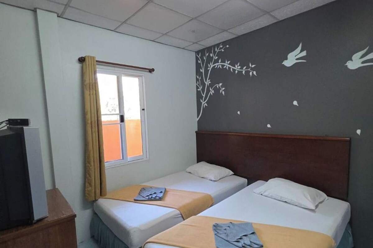 Une chambre d’hôtel avec deux lits, une fenêtre avec des rideaux, un mur décoré d’une fresque représentant un arbre et des oiseaux, et un bureau avec une télévision.