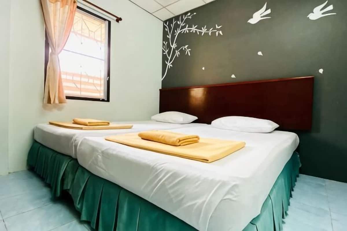 Een keurig opgemaakt bed met witte lakens en groene lakenkleed, een raam met gordijnen, en een muur met een muurschildering van een boom en een vogel.