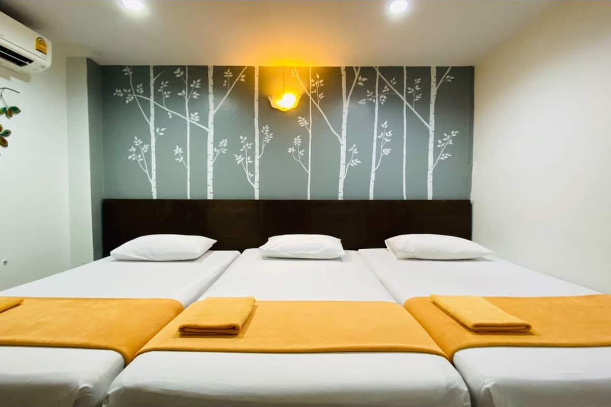 Hotelkamer met drie bedden, elk met witte lakens en een gele handdoek. Er is een wandairconditioner en een decoratieve muur met een boommotief.