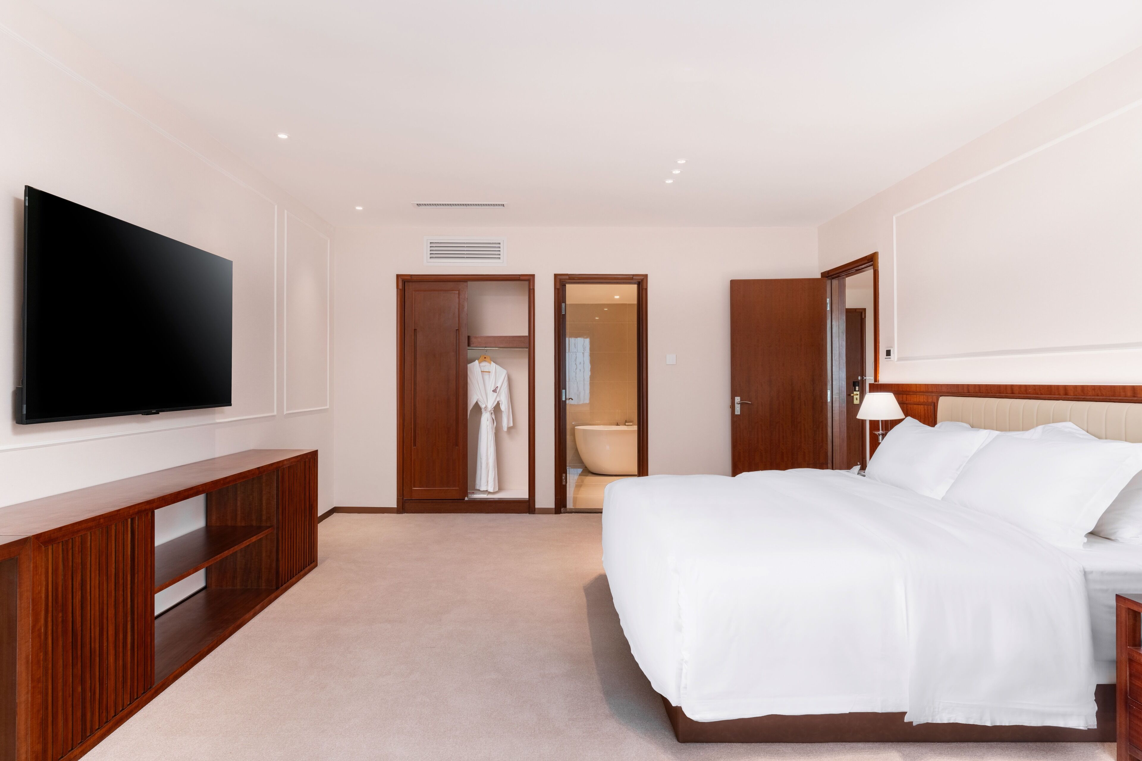 Quarto de hotel com uma televisão de tela plana grande fixada na parede, mesa consola de madeira, cama com roupa de cama branca, mesa de cabeceira com abajur e banheiro visível por uma porta aberta.