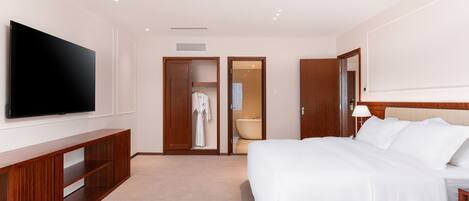 Quarto de hotel com uma televisão de tela plana grande fixada na parede, mesa consola de madeira, cama com roupa de cama branca, mesa de cabeceira com abajur e banheiro visível por uma porta aberta.