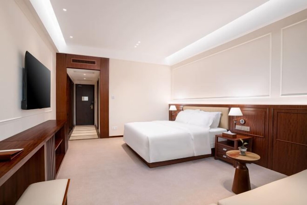 A hotel room with a bed, bedside table, lamp, and a TV mounted on the wall.
