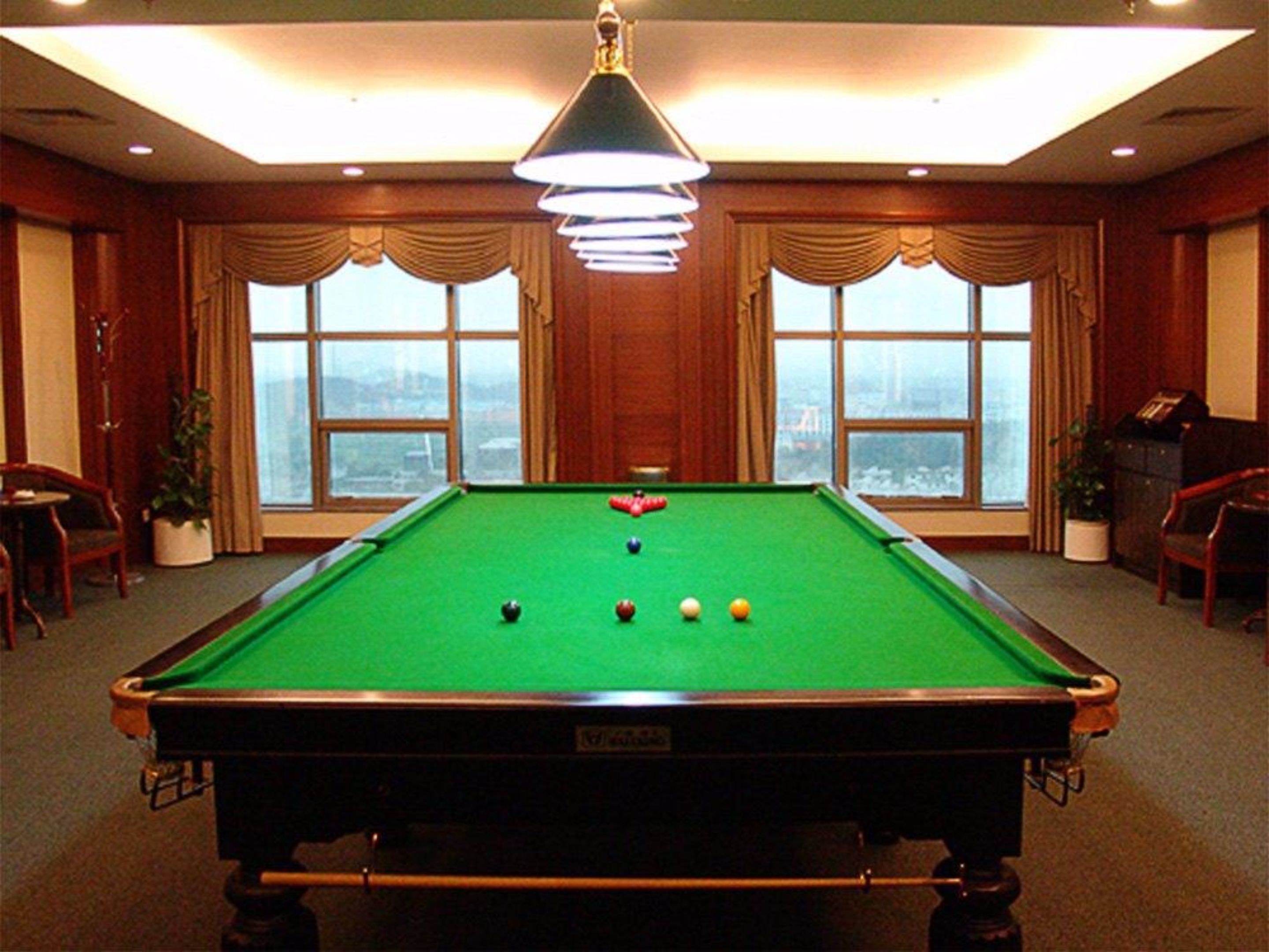 Une table de billard dans une pièce dotée de grandes fenêtres, offrant une vue sur le paysage urbain, et agrémentée d’un lustre.