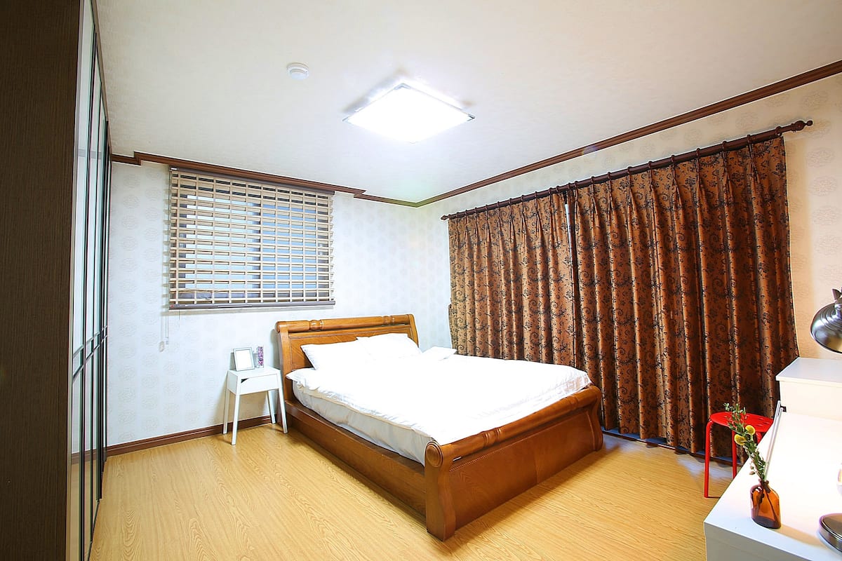 A bedroom with a wooden bed, a small bedside table, a desk with a lamp, and a window with blinds.