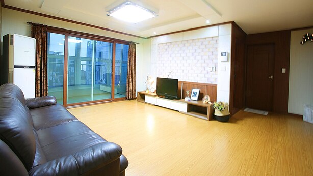 A living room with a brown leather sofa, a flat-screen TV on a wooden stand, and a sliding glass door leading to a balcony. - Woosung Hotel (Seogwipo)