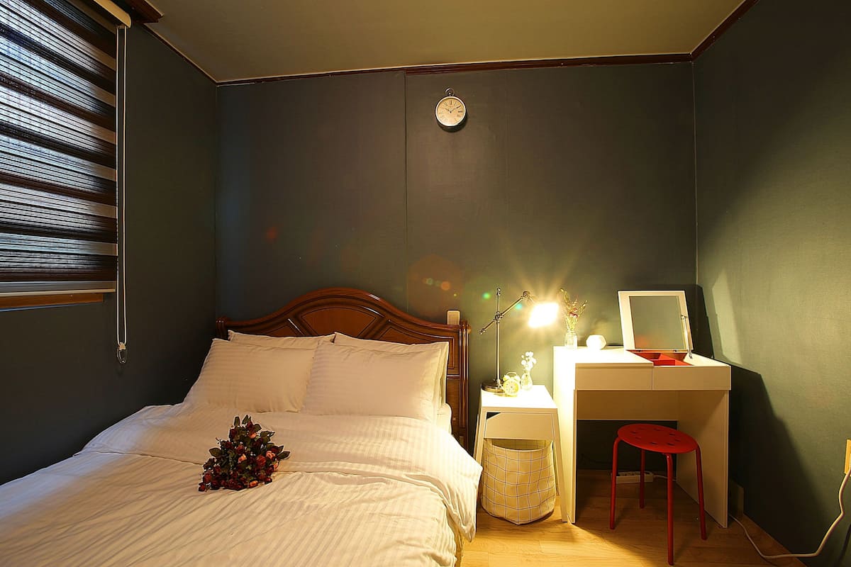 A bedroom with a bed, bedside table, desk, chair, and a clock on the wall.