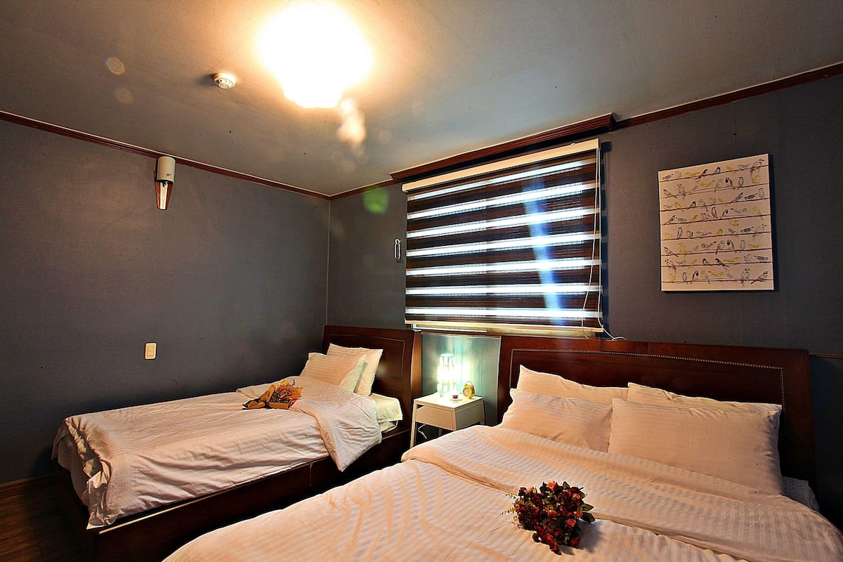A hotel room with two single beds, a nightstand with a lamp, a wall-mounted artwork, and a window with blinds.