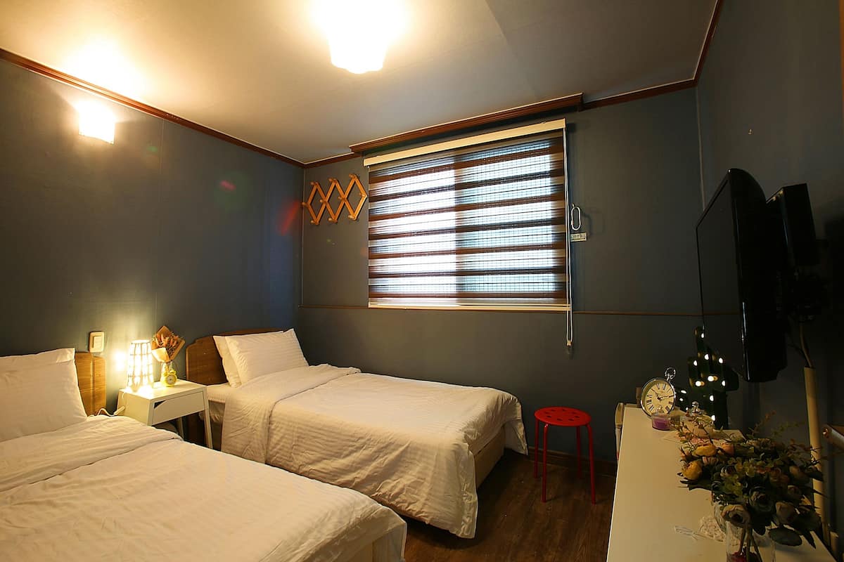 A hotel room with two beds, a nightstand with a lamp, a window with blinds, a red stool, and a wall-mounted TV.