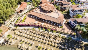 Vista aérea de un complejo turístico con varias villas frente al mar, un edificio central con techo de tejas y un amplio área de piscina con tumbonas y sombrillas.
