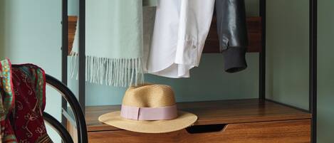 Una cama individual con estructura de madera, un pequeño escritorio con un sombrero y libros, y un perchero con chaquetas y una bufanda.