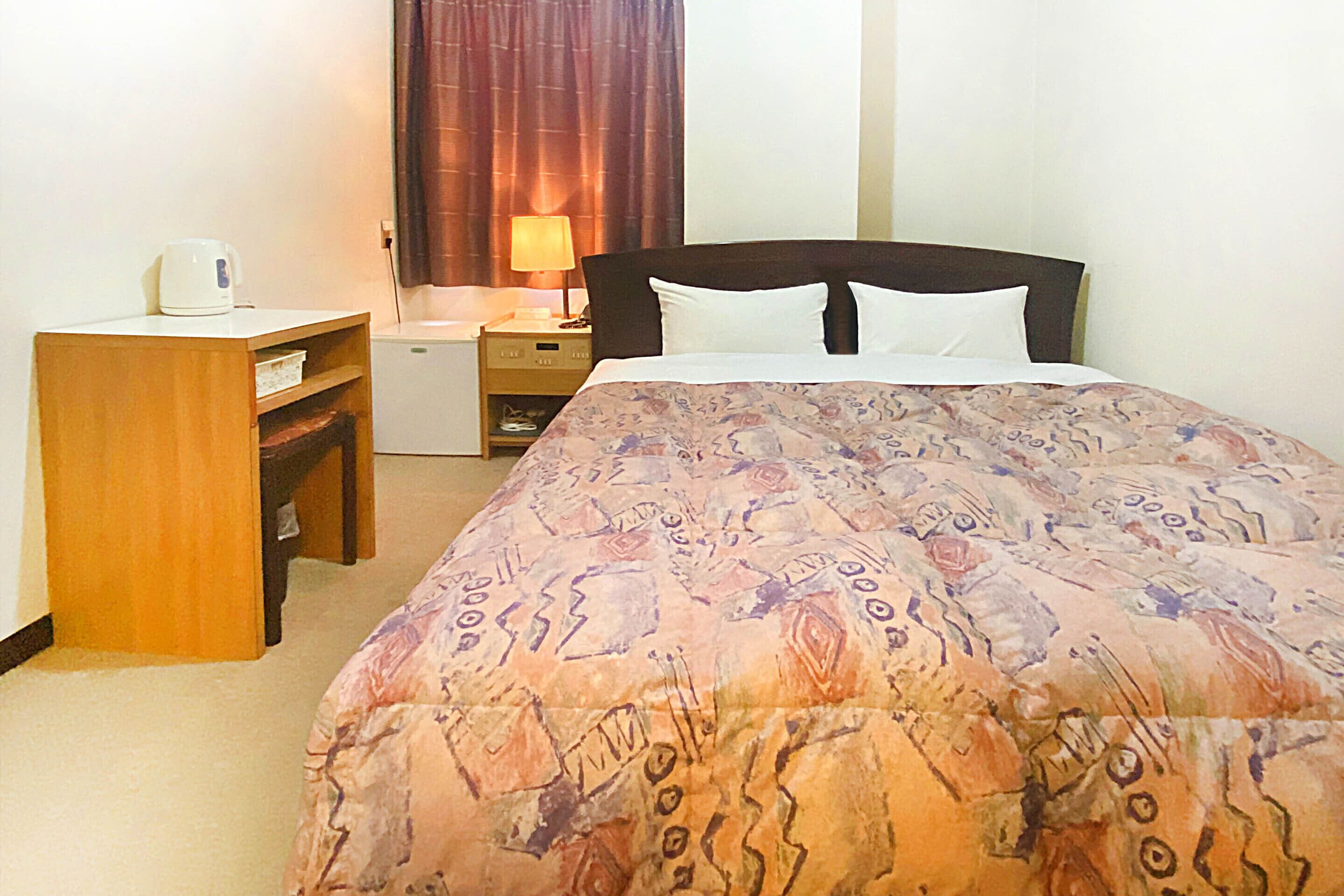 A hotel room with a bed, a desk, a nightstand, and a lamp.