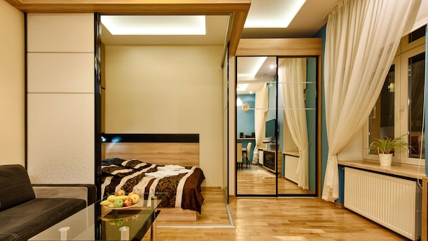 A modern hotel room with a bed, a glass table with fruits, a sofa, and a view into the adjacent room.
