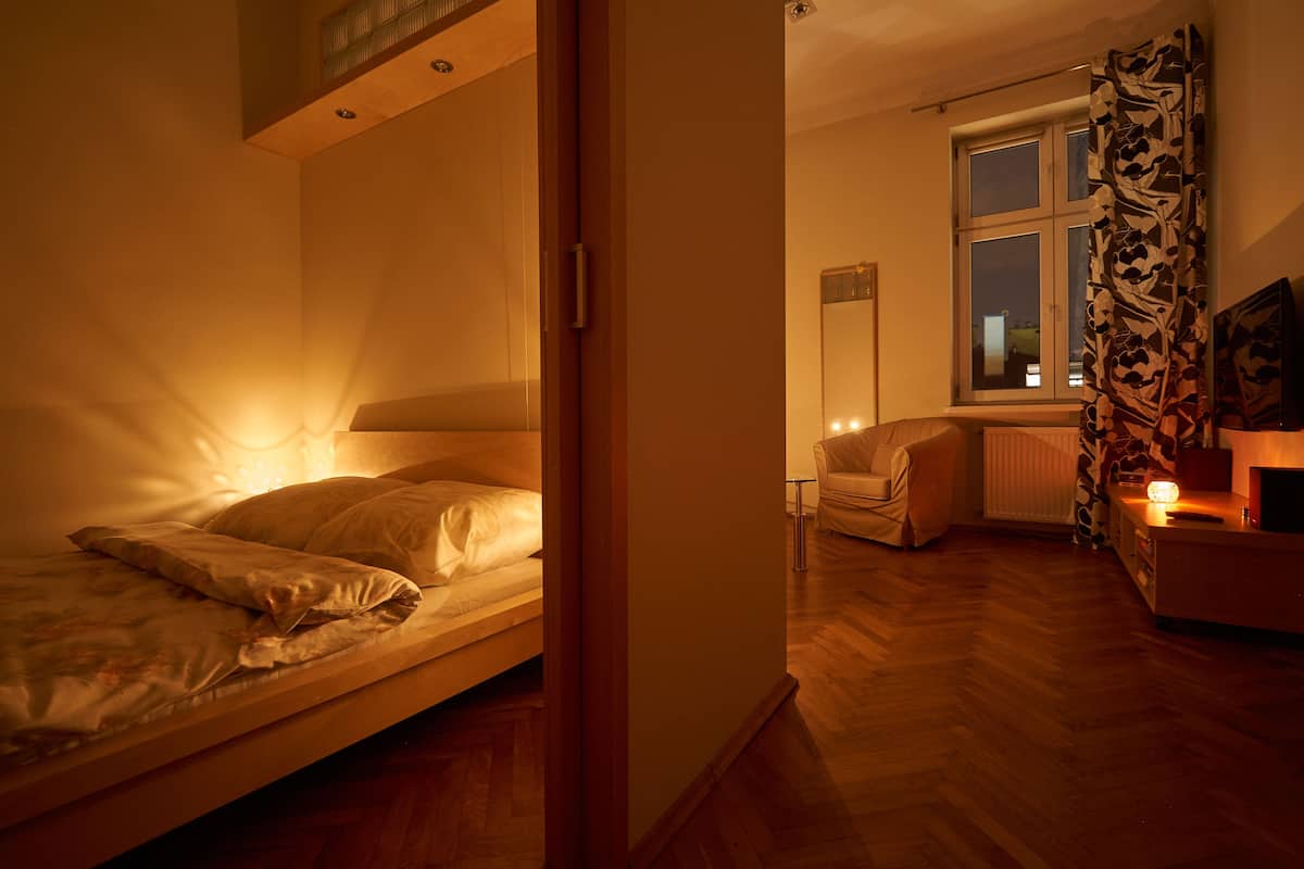 A bedroom with a single bed, a window with curtains, a radiator, and a small table with a lamp.
