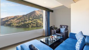 A modern living room with a large window offering a view of a lake and hills. - Via Lakhela Resort & Spa (Kumbhalgarh)