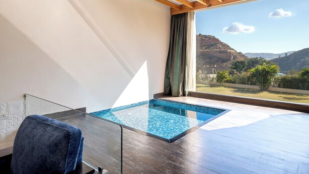 A modern room with a glass table, a chair, and a view of a swimming pool and mountains. - Via Lakhela Resort & Spa (Kumbhalgarh)