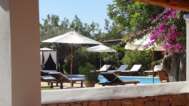 Zona de piscina con sillones de descanso y sombrillas, pared de piedra y vistas a la piscina y la vegetación.