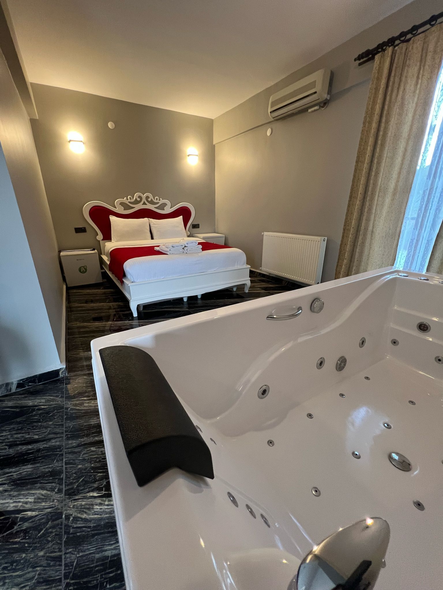Hotelkamer met een groot, wit bad, een bed met rode en witte beddengoed, een radiator en een raam met gordijnen.