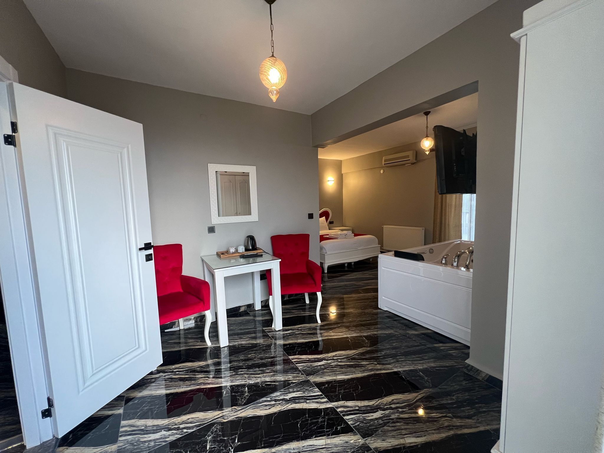 A hotel room with a marble floor, a white bed, a red chair, a small table with a kettle, and a bathroom with a white sink and mirror.
