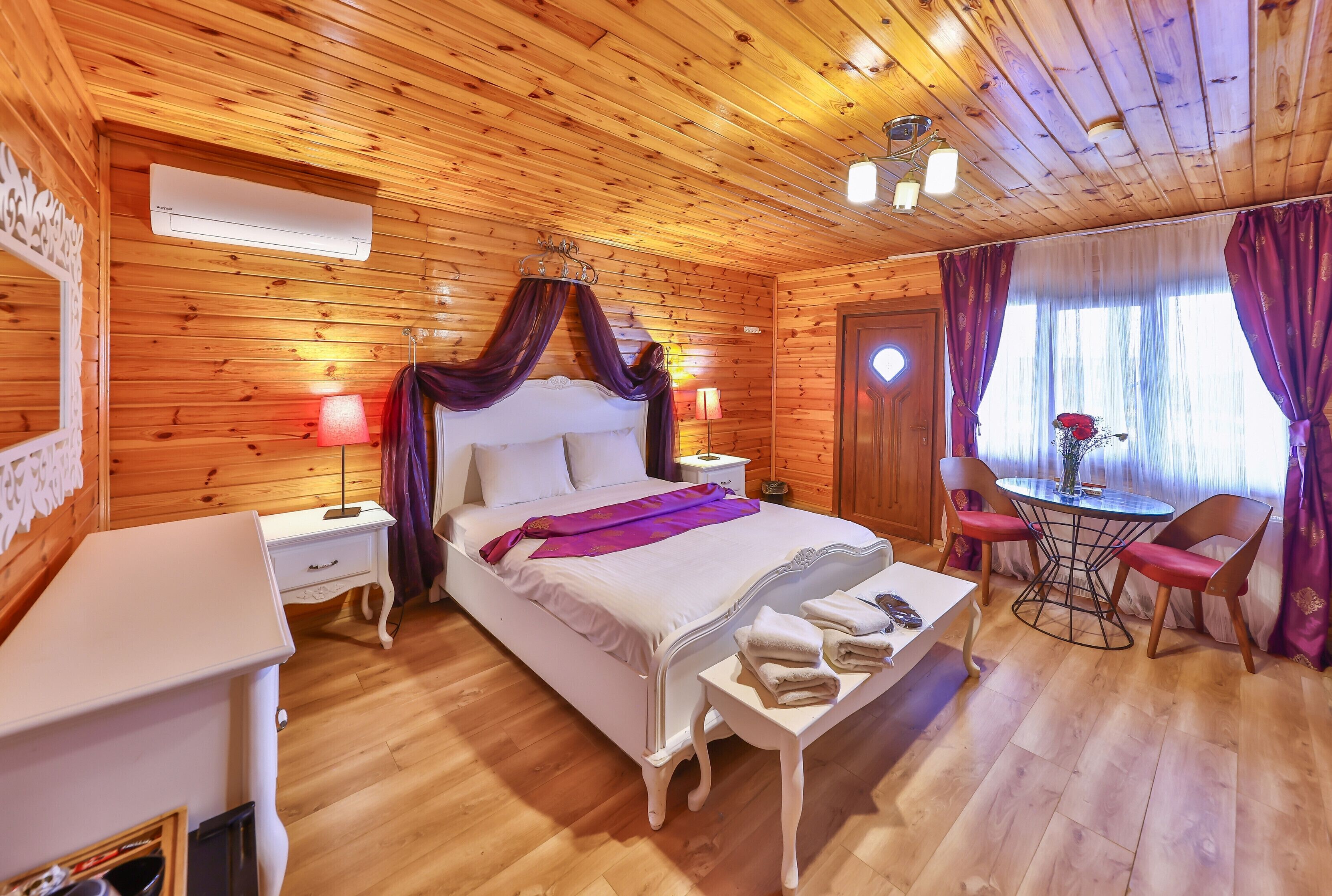 A wooden-paneled room with a bed, bedside tables, a table with chairs, and a window with curtains.