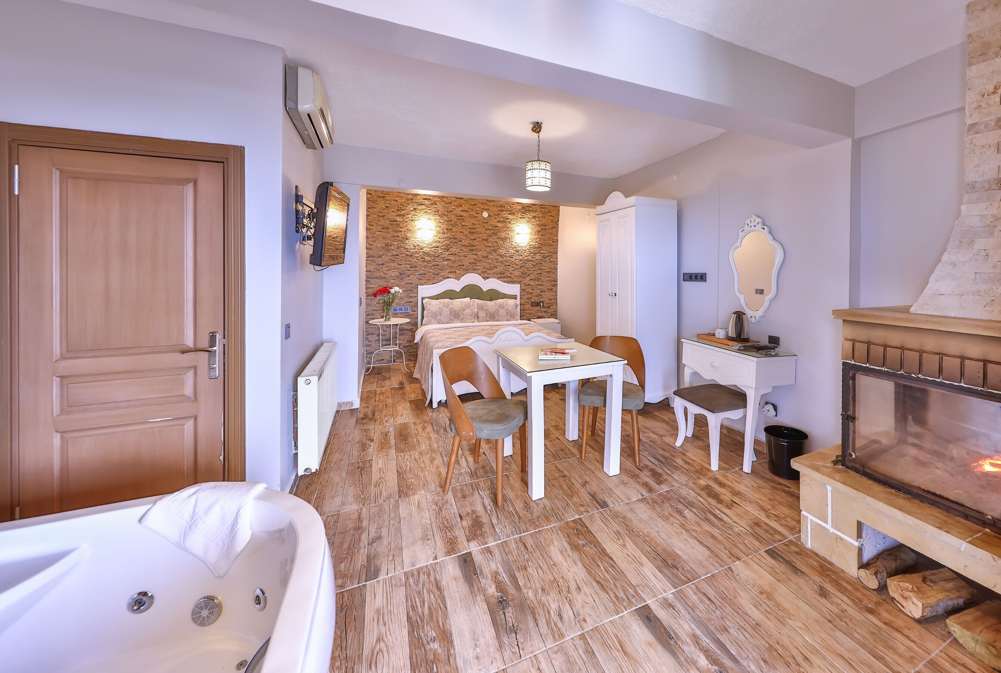 A bedroom with a wooden floor, a bed, a desk, a chair, a small table, a mirror, and a fireplace.