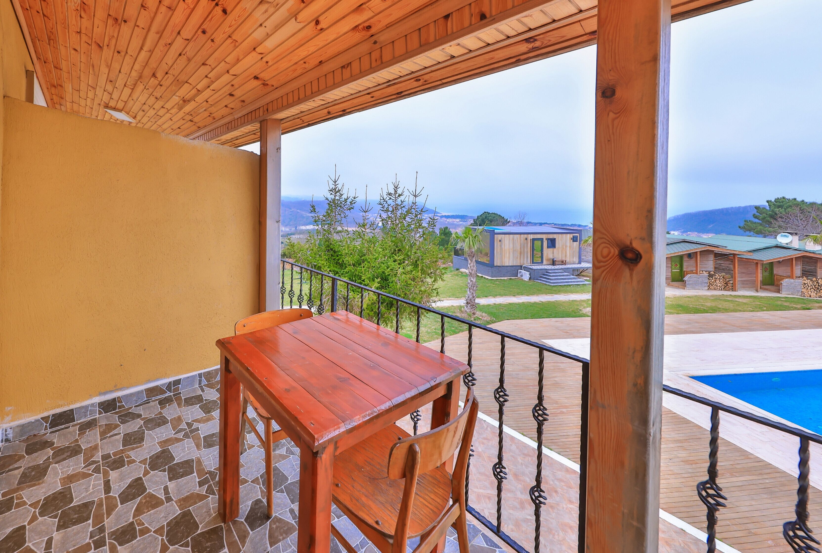 A wooden deck with a table and chairs, overlooking a pool and a view of buildings and trees.